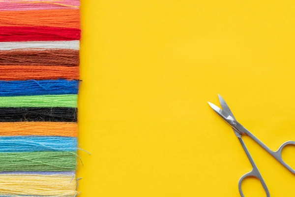 Multi-colored thread for embroidery laid out in a row on a yellow background and scissors for cutting. Top view, copy space, flat lay