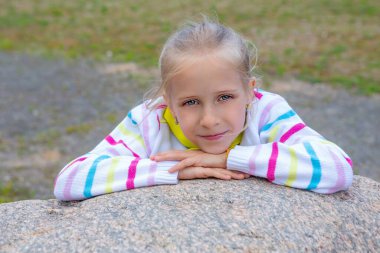 Dışarıda sarı uzun saçlı bir kızın portresi. Sarışın, uzun saçlı, beyaz bir kızın portresi. Sarı ceketli, sarı, mavi ve pembe çizgili sarı kravatlı kız kameraya bakıyor. Büyük taşın üzerinde oturan kız, onu...