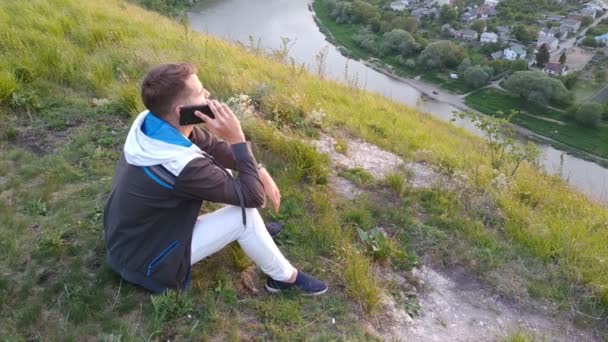 男は丘の中腹に座って電話で話します — ストック動画