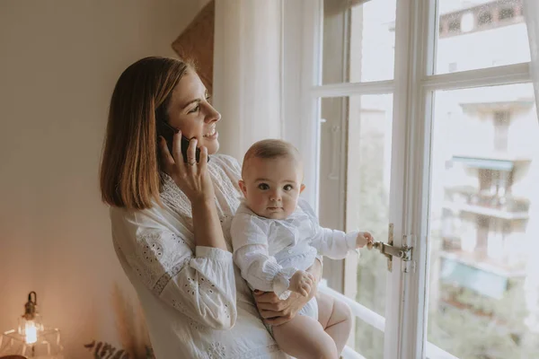 Young mother talks on the phone looking out the window with her baby in arms