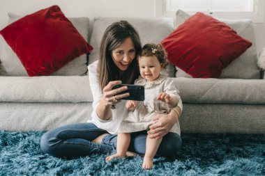 Genç ve güzel anne, kızıyla oturma odasında yerde telefonla oynuyor.