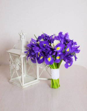 Bir buket iris. Masanın üzerinde mor-mavi renkli, beyaz güllü pofuduk bir gelin buketi duruyor. Masanın üzerinde dekoratif bir fener var..