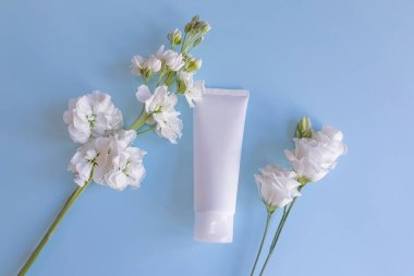 Beyaz sıkılmış plastik tüp ve soluk mavi arka planda beyaz çiçeklerin üst görüntüsü. İlaç veya kozmetik için beyaz parlak plastik tüp - krem, jöle, cilt bakımı, diş macunu.