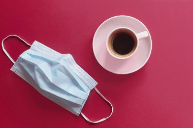 Bir fincan kahve ve çıkarılmış tıbbi maske. Kahvaltını kafede yap. Modern gerçeklik ve yaşam konsepti. Mavi cerrahi koruyucu maske ve vişneli arka planda pembe bardak.. 
