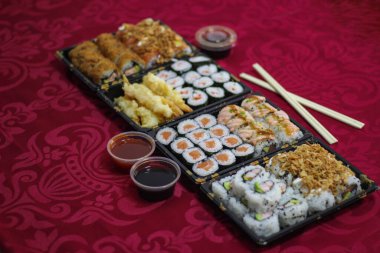 İçinde somon balığı, Hosomaki, California, UrAMAKI, kızarmış soğan, kızarmış soğan, karides, tempura karidesi, bir kase acı tatlı ve ekşi sos, kırmızı masa örtüsü üzerinde çubuklar bulunan 4 Sushi paket servis kutusu.