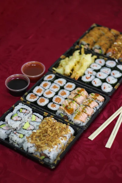 4 Sushi take-away delivery boxes with salmon Hosomaki, Californias, URAMAKI, fried onion, crispy onion, breaded shrimp, tempura shrimp, bowl of bittersweet, sweet and sour sauce, chopsticks on a red t