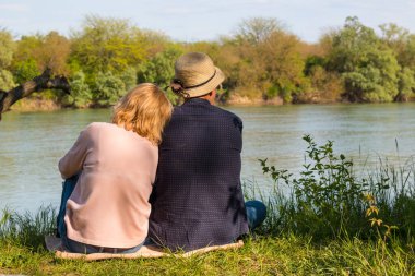 Birbirine aşık bir çift nehir kıyısındaki yeşil çimlerin üzerinde oturuyor. Günlük kıyafetlerle. Bahar. Fotoğraf, arkadan çekilmişti. Yatay düzenleme.