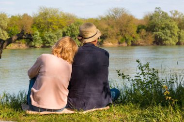 Birbirine aşık bir çift nehir kıyısındaki yeşil çimlerin üzerinde oturuyor. Günlük kıyafetlerle. Bahar. Fotoğraf, arkadan çekilmişti. Yatay düzenleme.