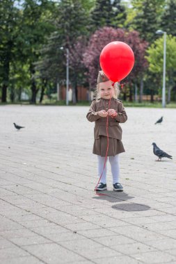 Askeri üniforma giymiş küçük bir kız. Elinde büyük kırmızı bir topla meydanda yürür. Zafer töreninden önce. Ciddi bir ifade. Bahar. Gün