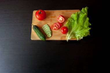 Siyah bir masada bambu doğrama tahtasında dilimlenmiş salatalık, domates ve marul yaprağı. Yatay çerçeve