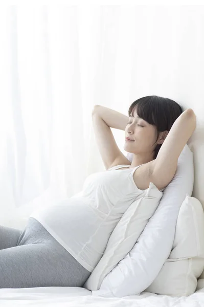 若いです妊娠中のアジアの女性座っていますベッドで彼女の手後ろ頭 — ストック写真