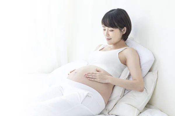 若いです妊娠中のアジアの女性座っていますベッドに彼女の手でザ — ストック写真