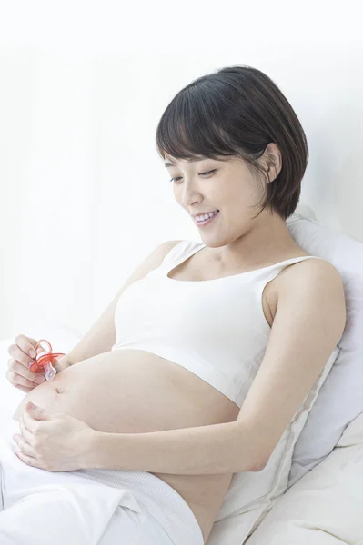 若い妊娠中のアジアの女性がベッドに座って赤ちゃんのペースメーカーを持って — ストック写真