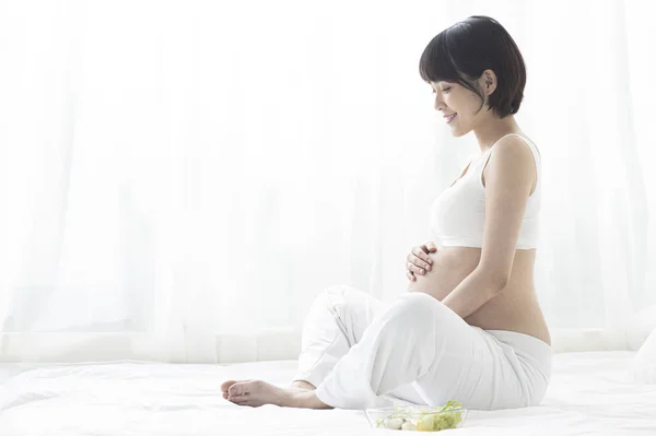 若いです妊娠中のアジアの女性は ハスの位置にベッドの上に座って 彼女の腹にしがみつく — ストック写真