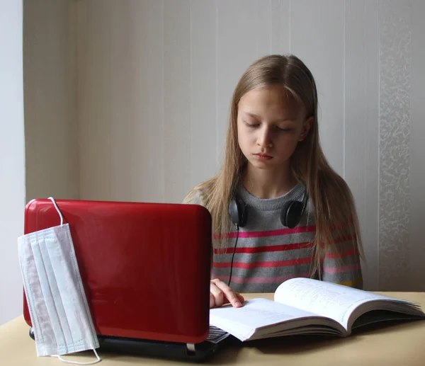 A blonde preteen girl with headphones sits at a table with a computer and a book. Home schooling. The schools are closed on quarantine due to the pandemic of coronavirus Covid-19. Distance education