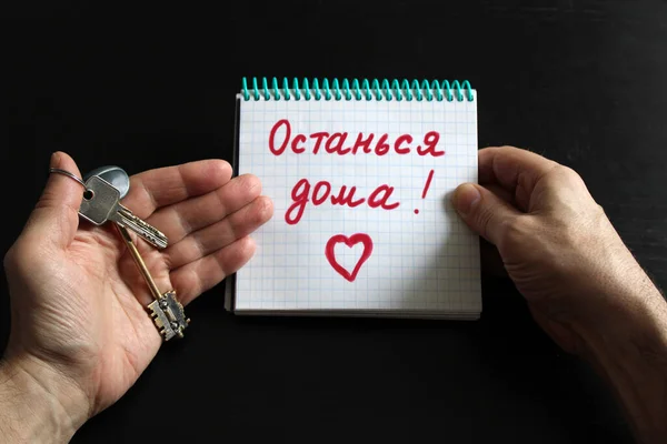 Erkek eli evin anahtarlarını ve üzerinde not olan bir defter tutuyor. Evde Rusça konuş. Koronavirüs Covid-19 salgınından dolayı karantina ya da kendini izole etme. Ev tabelasında kal.