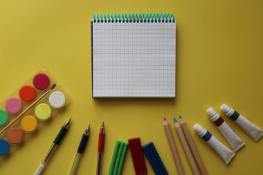 Okul kırtasiyesinin üst görüntüsü: boyalar, renkli kalemler, kalemler, plastik kalemler, not defteri ve sarı arka plandaki elma. Okula geri döndüm. Metnin için boşluk kopyala