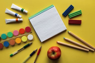 Okul kırtasiyesinin üst görüntüsü: boyalar, renkli kalemler, kalemler, plastik kalemler, not defteri ve sarı arka plandaki elma. Okula geri döndüm. Metnin için boşluk kopyala