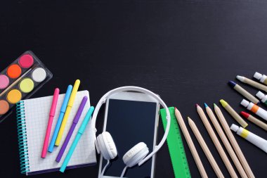 Okul kırtasiyesinin en üst görüntüsü: siyah kalemler, boyalar, pastel kalemler, işaretler, cetvel, not defteri, elektronik tablet ve siyah ahşap masa üzerinde kulaklıklar. Okula geri döndüm. Boşluğu kopyala