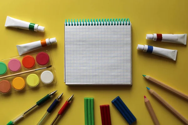 Okul kırtasiyesinin üst görüntüsü: boyalar, renkli kalemler, kalemler, plastik kalemler, not defteri ve sarı arka plandaki elma. Okula geri döndüm. Metnin için boşluk kopyala
