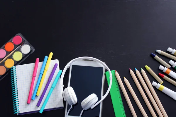 Okul kırtasiyesinin en üst görüntüsü: siyah kalemler, boyalar, pastel kalemler, işaretler, cetvel, not defteri, elektronik tablet ve siyah ahşap masa üzerinde kulaklıklar. Okula geri döndüm. Boşluğu kopyala