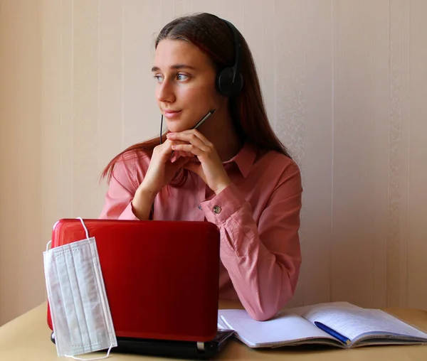 Kulaklıklı bir kız hayal kurarak masada bilgisayar ve kitaplarla oturuyor. Evde koruyucu maskeye ihtiyacı yok. Coronavirus Covid-19 salgını nedeniyle okullar ve üniversiteler karantinaya alınmıştır. Uzaklık eğitimi içeriği