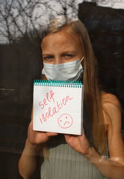 Daire penceresinin arkasında yüzü koruyucu maskeli sarışın bir kızın bulanık portresi üzerinde 