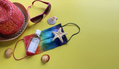 Yaz tatilinde gökkuşağı renklerine boyanmış koruyucu maske, antiseptik, plaj şapkası, güneş gözlüğü, deniz kabuğu ve sarı arka planda deniz yıldızı ile yatıyordu. Seyahat ve Coronavirus covid-19. Boşluğu kopyala