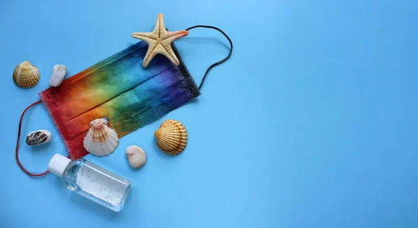 Yaz tatilinde gökkuşağı renklerine boyanmış koruyucu maske, antiseptik, deniz kabuğu ve mavi arka planda deniz yıldızı ile yatıyordu. Coronavirus covid-19 ve diğer enfeksiyonlara karşı seyahat ve koruma. Boşluğu kopyala