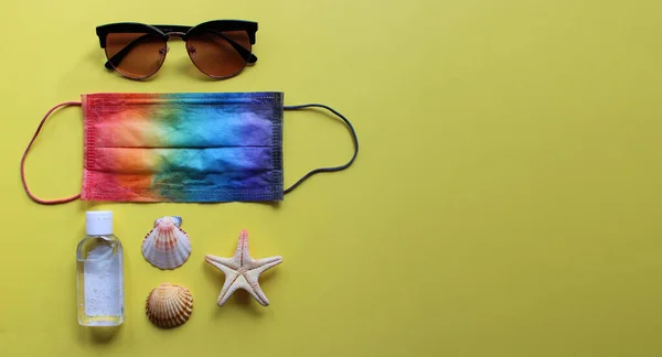 Yaz tatilinde gökkuşağı renklerine boyanmış koruyucu maske, antiseptik, deniz kabuğu ve güneş tarafından aydınlatılan mavi arka planda deniz yıldızı ile yatıyordu. Coronavirus covid-19 ve diğer enfeksiyonlara karşı seyahat ve koruma.