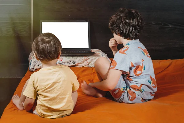 Children sit on an orange bed and watch cartoons on a laptop