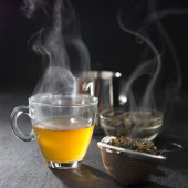 Proces vaření čaje, čajový obřad, šálek čerstvě uvařeného žlutého nebo zeleného bylinkového čaje, teplé měkké světlo, tmavá nálada.Teplá voda se nalévá z konvice do šálku s čajovými lístky.