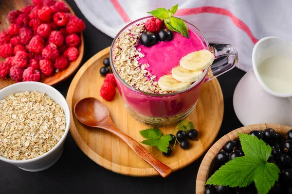 Healthy breakfast,fitness food for diet, Raspberry and currant berry bowl smoothie in a one glass cup with a wooden spoon,mint leaf, oatmeal in white dish on a dark background.