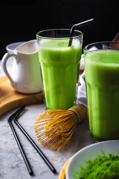 Matcha green tea powder latte with milk in two high glass on stone table, black straw, wooden plate and tray,bamboo tasaku and whisk,dark backdrop