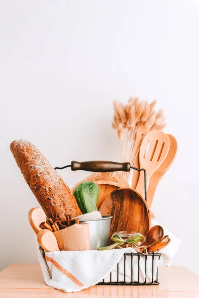 Gift basket with kitchen accessories and utensils made of wood and bamboo, bread, spices, fresh lettuce leaves, cotton towel on the table against a white wall in a bright room.