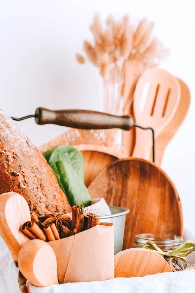 Gift basket with kitchen accessories and utensils made of wood and bamboo, bread, spices, fresh lettuce leaves, cotton towel on the table against a white wall in a bright room.
