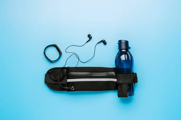 Running belt, fitness smart bracelet, water bottle and headphones on a trendy blue background,space for text