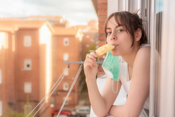 Young girl with face mask leaning out of the window eating ice cream during quarantine. Person confined in summer eating ice cream