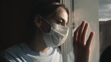 Karantina sırasında maskeli hasta kız pencereden dışarı bakıyor. Üzgün bir kadın sokağa çıkmak istiyor.