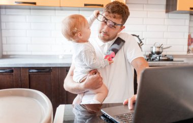 Evde dizüstü bilgisayarı ve elinde bebekle çalışan yakışıklı bir genç. Evde kal konsepti. Çocuklu ev ofisi. 