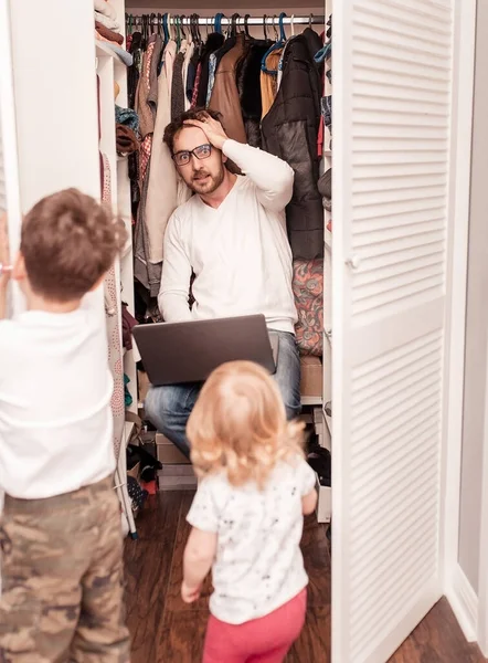 Busy father trying to find a quiet place in a wardrobe  to hide from children for a work at the laptop. Stay home concept. Quarantine. Freelancer.