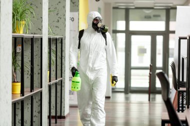 Koruyucu ekipmanlı bir adam ofiste spreyle dezenfekte ediyor. Coronavirus covid-19 hastalığından dolayı yüzey tedavisi. Beyaz takım elbiseli bir adam odayı sprey silahla dezenfekte ediyor. Virüs salgını