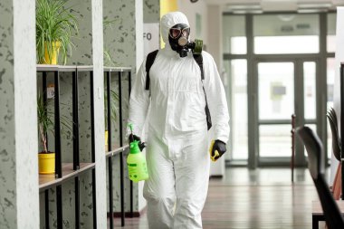 Koruyucu ekipmanlı bir adam ofiste spreyle dezenfekte ediyor. Coronavirus covid-19 hastalığından dolayı yüzey tedavisi. Beyaz takım elbiseli bir adam odayı sprey silahla dezenfekte ediyor. Virüs salgını