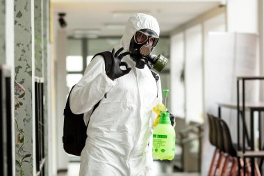 Koruyucu ekipmanlı bir adam ofiste spreyle dezenfekte ediyor. Coronavirus covid-19 hastalığından dolayı yüzey tedavisi. Beyaz takım elbiseli bir adam odayı sprey silahla dezenfekte ediyor. Virüs salgını