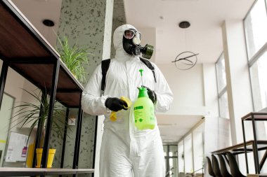 Koruyucu ekipmanlı bir adam ofiste spreyle dezenfekte ediyor. Coronavirus covid-19 hastalığından dolayı yüzey tedavisi. Beyaz takım elbiseli bir adam odayı sprey silahla dezenfekte ediyor. Virüs salgını