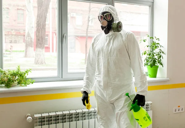 A man in protective equipment disinfects with a sprayer in the office. Surface treatment due to coronavirus covid-19 disease. A man in a white suit disinfects the room with a spray gun. Virus pandemic