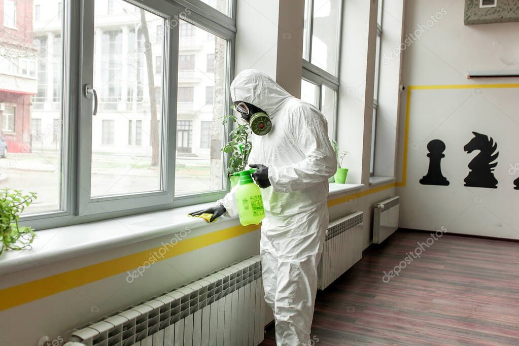 A man in protective equipment disinfects with a sprayer in the office. Surface treatment due to coronavirus covid-19 disease. A man in a white suit disinfects the room with a spray gun. Virus pandemic