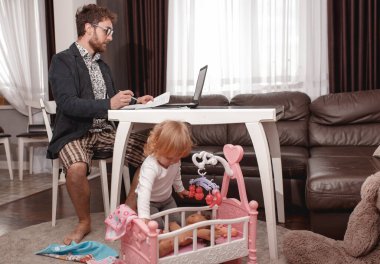 Genç bir iş adamı evden laptopta video konferansı yapıyor. Güçlü bir ceket, gömlek ve şort giyiyor. Küçük kız babasının yanında oturuyor ve bebeklerle oynuyor..