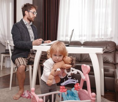 Genç bir iş adamı evden laptopta video konferansı yapıyor. Güçlü bir ceket, gömlek ve şort giyiyor. Küçük kız babasının yanında oturuyor ve bebeklerle oynuyor..