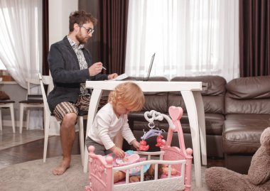 Genç bir iş adamı evden laptopta video konferansı yapıyor. Güçlü bir ceket, gömlek ve şort giyiyor. Küçük kız babasının yanında oturuyor ve bebeklerle oynuyor..